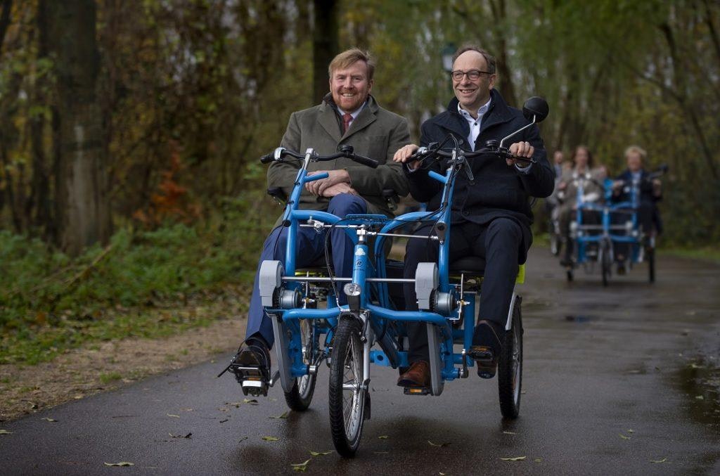 Koning fietst op duofiets met oprichter van stichting Fietsmaatjes